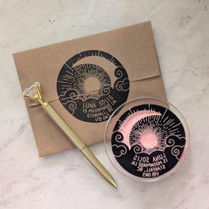 May include: A gold pen with a diamond-shaped top rests beside a brown envelope and a clear, round container. The envelope and container feature a black and pink design with the text "LUNA SOLIS 13 MOONPHASE LN. STARFALL, BC V1B 0N3".