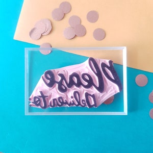 May include: A clear acrylic stamp with a pink rubber surface. The stamp reads "Please deliver to" in a black cursive font.