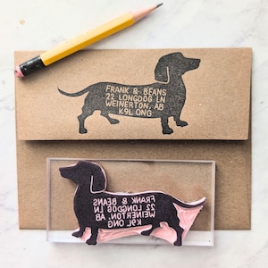 Könnte beinhalten: Ein schwarzer Dackel-Stempel mit dem Text "FRANK & BEANS 22 LONGDOG LN WEINERTON, AB K9L ONG" auf einem braunen Umschlag. Ein gelber Bleistift liegt auf dem Umschlag. Der Stempel ist auf einem Acrylblock.