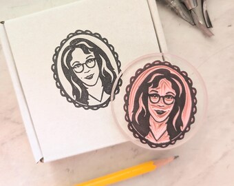 Pode incluir: Uma caixa de papelão branca com um carimbo de tinta preta de uma pessoa de óculos dentro de uma moldura oval festonada. Ao lado, há um carimbo de borracha rosa circular com o mesmo retrato, ferramentas de entalhe e um lápis amarelo.