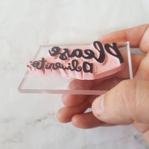 May include: A pink rubber stamp with the text "Please don't be a trouble" in black lettering. The stamp is mounted on a clear acrylic base.
