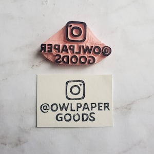 May include: A pink rubber stamp with the text "GOODS OWLPAPER" and an Instagram logo. The stamp is on a white surface. Below the stamp is a white card with the text "@OWLPAPER GOODS" and an Instagram logo.