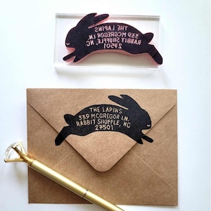 May include: A clear acrylic rubber stamp shaped like a rabbit with the text THE LAPINS 339 MCGREGOR LN. RABBIT SHUFFLE, NC 27501, a brown paper envelope with the same rabbit design stamped in black ink, and a gold pen with a clear crystal topper.