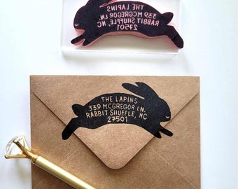 Pode incluir: Um carimbo de borracha acrílico transparente em forma de coelho com o texto THE LAPINS 339 MCGREGOR LN. RABBIT SHUFFLE, NC 27501, um envelope de papel castanho com o mesmo design de coelho estampado a preto e uma caneta dourada com cristal.