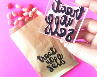 May include: A brown paper bag with the text "treat yo self" printed in black. A clear acrylic stamp with the text "treat yo self" in black lettering is held in a hand.