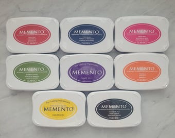 Pode incluir: Conjunto de dez almofadas de tinta Memento em várias cores, incluindo vermelho, azul, rosa, verde, roxo, laranja, amarelo e preto. Cada almofada de tinta apresenta o logotipo Memento e o nome da cor da tinta.
