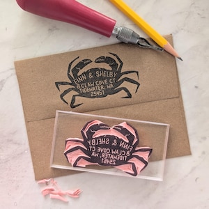 May include: A crab-shaped rubber stamp with the text "FINN & SHELBY 8 CLAW COVE CT. TIDEWATER, WA 23451" printed on a brown envelope. A pink carving tool and a yellow pencil are also in the image.