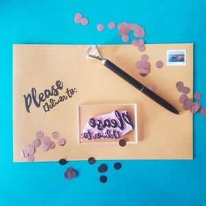 May include: A brown envelope with the text "Please deliver to:" written in black ink. A clear acrylic stamp with the text "Please deliver to:" in black ink is on the envelope. The envelope is surrounded by small, pink, metallic confetti.