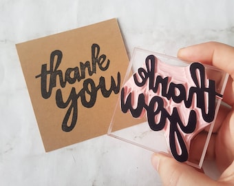 May include: A clear acrylic stamp with a pink rubber surface. The stamp reads "thank you" in a black, handwritten font.