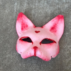 May include: Pink felt cat mask with black eyes and a red heart on the forehead.