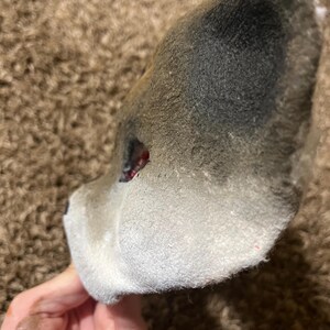 May include: A gray and white wolf mask, with a dark gray top and white bottom. The mask has a red eye and is held by a hand.
