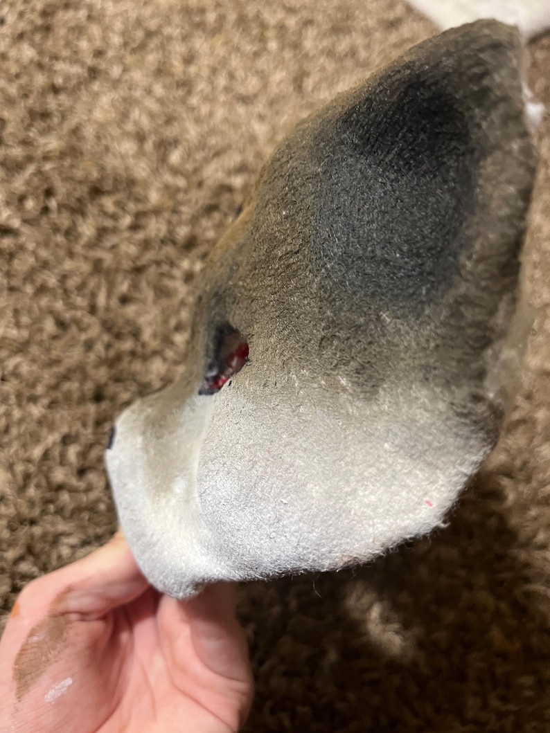 May include: A gray and white wolf mask, with a dark gray top and white bottom. The mask has a red eye and is held by a hand.