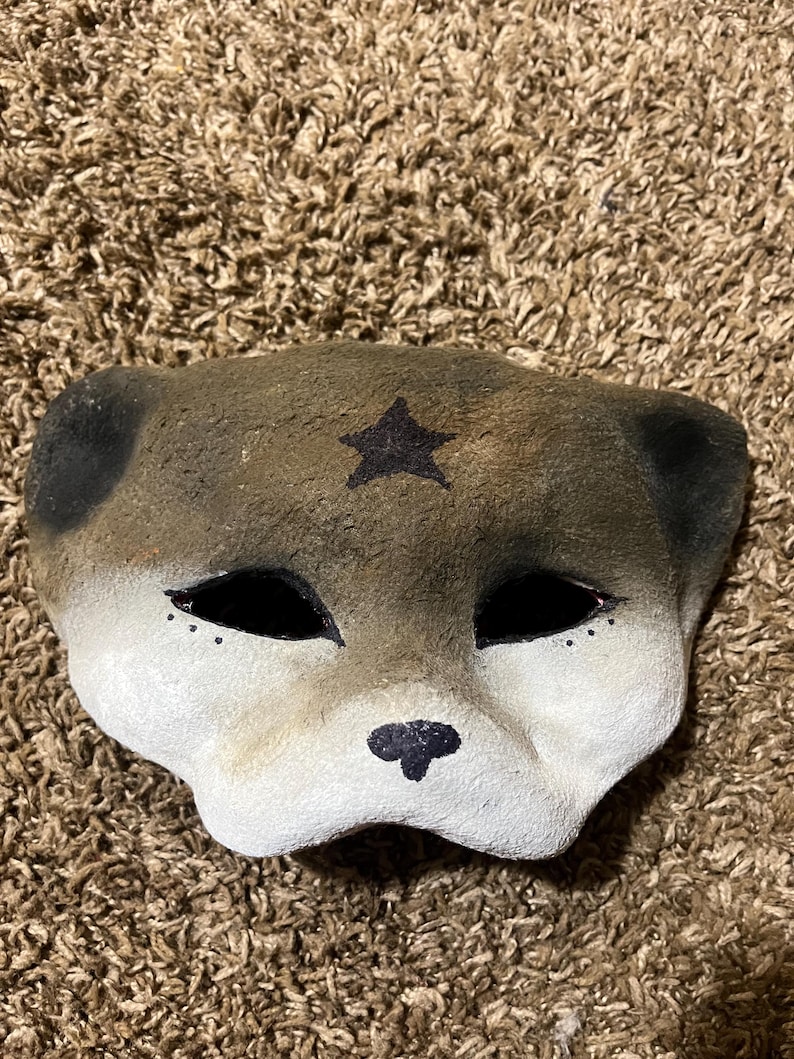 May include: A handmade mask in the shape of a dog's face. The mask is primarily gray and white, with black accents for the eyes, nose, and ears. A black star is painted on the forehead. The mask is likely for costume or decorative purposes.