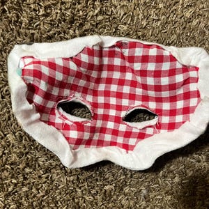 May include: A red and white checkered fabric mask with eye holes, trimmed with white fabric. The mask is designed to be worn on the face. The mask is likely for a costume or novelty item.
