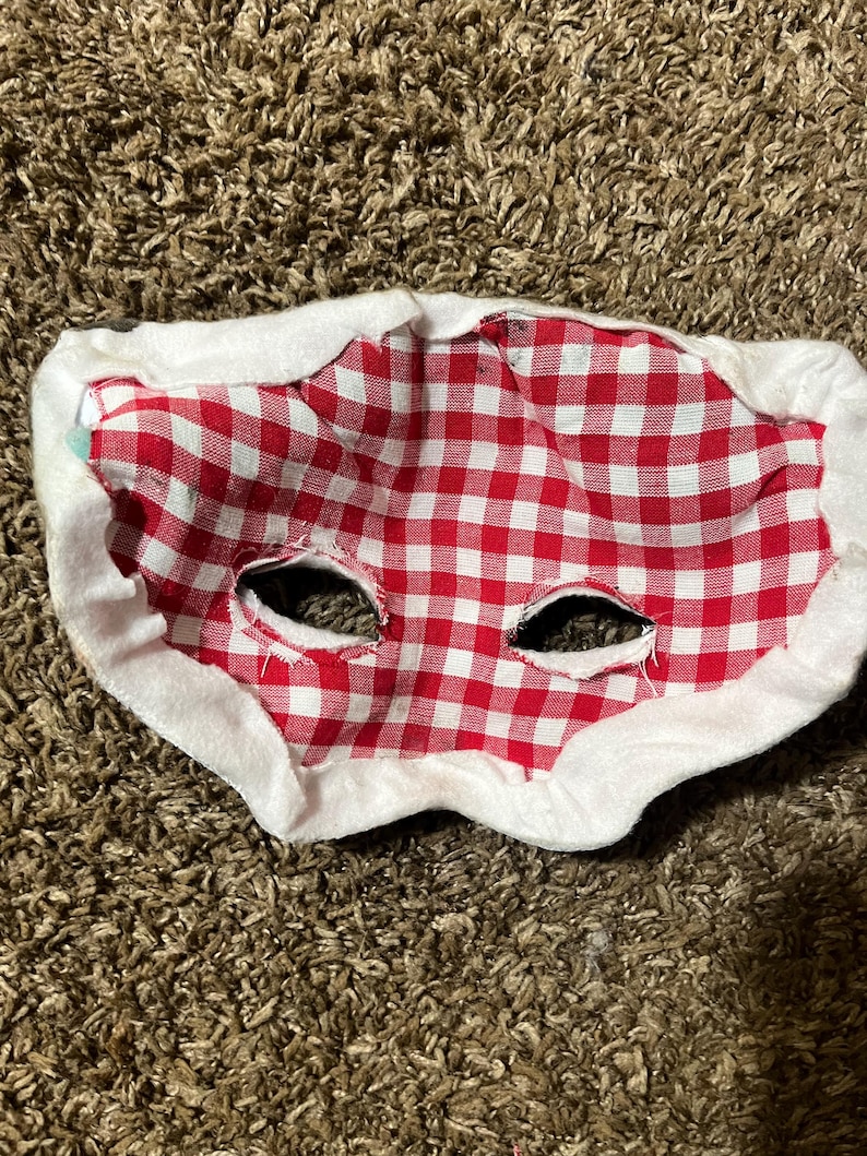 May include: A red and white checkered fabric mask with eye holes, trimmed with white fabric. The mask is designed to be worn on the face. The mask is likely for a costume or novelty item.