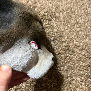 May include: A handmade, gray and brown wolf mask with a black nose and a red and white eye. The mask is made of a textured material and is partially visible.