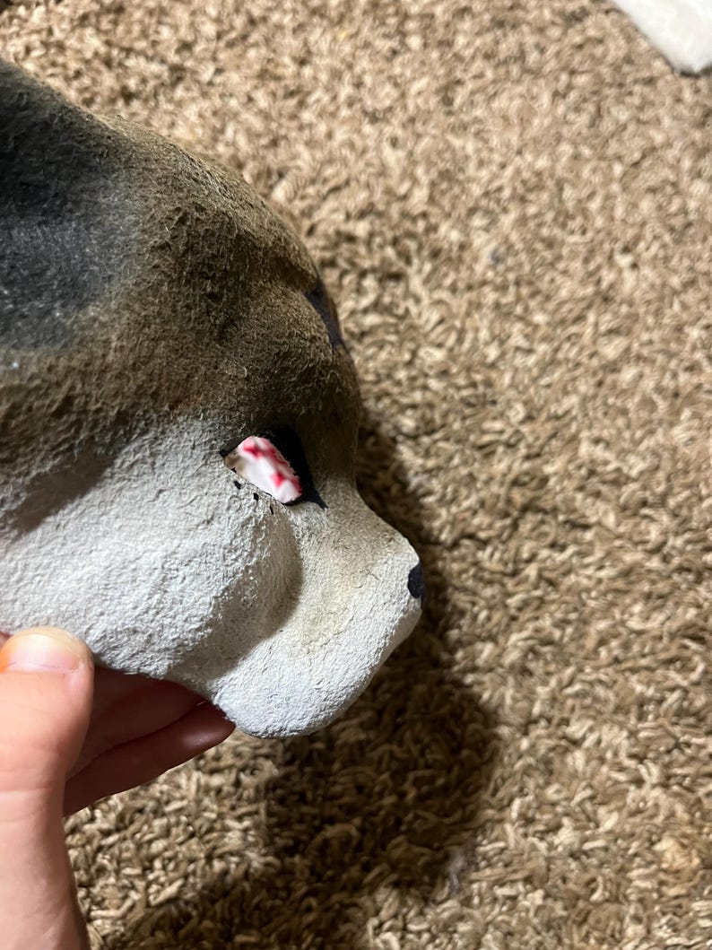 May include: A handmade, gray and brown wolf mask with a black nose and a red and white eye. The mask is made of a textured material and is partially visible.