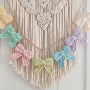 Puede incluir: Una guirnalda de lazos de ganchillo en colores pastel: amarillo, verde menta, rosa, verde claro, azul y lavanda. Los lazos están ensartados en una cuerda, frente a un tapiz de macramé.