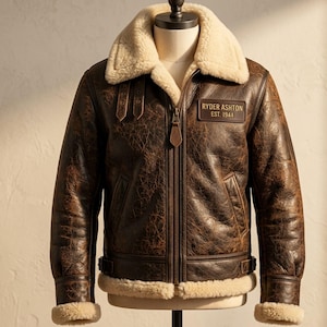 Pode incluir: Um casaco de couro envelhecido castanho-escuro com gola e forro de pele de carneiro cor de creme. O casaco apresenta um fecho central, dois bolsos laterais, tiras laterais ajustáveis e um emblema retangular com RYDER ASHTON EST. 1944.