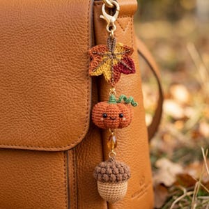Può includere: Un ciondolo all'uncinetto con motivi autunnali. Presenta una foglia d'acero, una zucca sorridente e una ghianda, nei toni dell'arancione, giallo e marrone. Il ciondolo è attaccato a un fermaglio color oro, ideale per una borsa o un portachiavi.