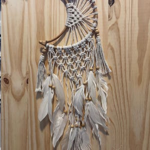 May include: A handcrafted dream catcher in a crescent moon shape. The dream catcher features a woven web design with white feathers and wooden beads. The frame is made of a curved wooden branch. The dream catcher is hanging against a wooden background.