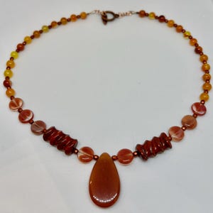 May include: A handcrafted necklace featuring a variety of orange and yellow beads. The necklace includes a teardrop-shaped pendant and dark red, cylindrical beads. The clasp is copper-colored.