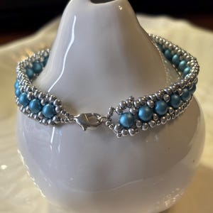 以下が含まれることがあります： シルバーカラーのビーズと水色のビーズを使ったビーズブレスレット。白い陶器の花瓶に飾られています。ブレスレットにはシルバーカラーの留め金が付いています。