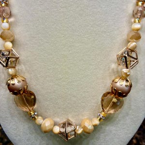 Peut inclure: Un collier de perles aux formes et couleurs variées. Il présente des formes géométriques dorées, des perles rondes et des perles facettées dans des tons crème, or et ambre. Le collier est présenté sur un fond neutre.