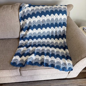 May include: A blue and white crocheted blanket with a wave pattern. The blanket is folded and placed on a light brown couch.