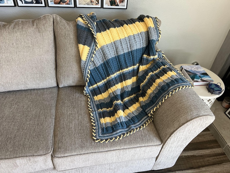 May include: A striped crocheted throw blanket in shades of blue, gray, and yellow with a crocheted border. The blanket is draped over a gray couch.