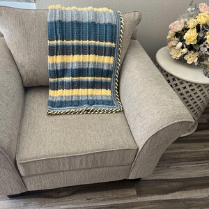 May include: A gray armchair with a yellow, blue, and gray striped crocheted throw blanket on top. The armchair is in a room with a wooden floor and a small white side table with a bouquet of flowers.