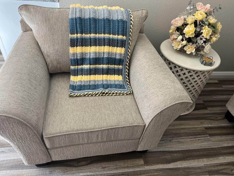 May include: A gray armchair with a yellow, blue, and gray striped crocheted throw blanket on top. The armchair is in a room with a wooden floor and a small white side table with a bouquet of flowers.