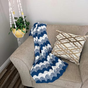 May include: A blue and white crocheted throw blanket with a wave pattern is draped over a beige armchair. A brown and beige patterned pillow is on the chair.