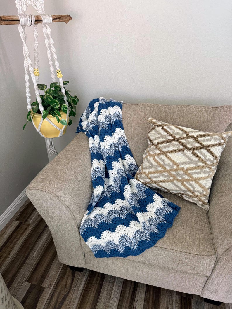 May include: A blue and white crocheted throw blanket with a wave pattern is draped over a beige armchair. A brown and beige patterned pillow is on the chair.