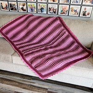 May include: A crocheted blanket in shades of pink and purple with a scalloped edge. The blanket is draped over a couch.