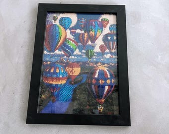 May include: A framed diamond painting depicting a colorful scene of hot air balloons floating in the sky. The balloons are various shades of blue, green, red, yellow, and purple. The background is a light blue sky with white clouds.