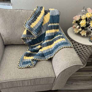 May include: A striped crocheted throw blanket in shades of blue, yellow, and gray with a yellow crocheted border. The blanket is draped over a gray armchair.