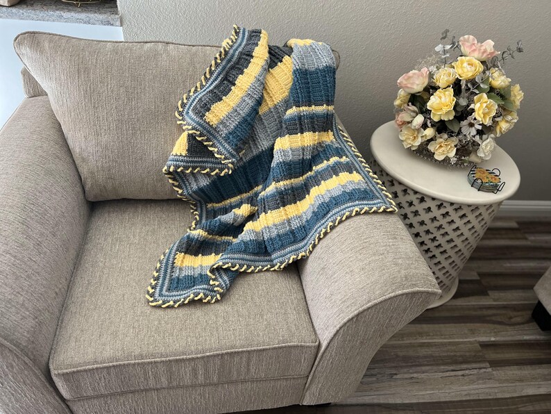 May include: A striped crocheted throw blanket in shades of blue, yellow, and gray with a yellow crocheted border. The blanket is draped over a gray armchair.