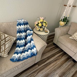 May include: Two beige armchairs with throw pillows, one with a blue and white crocheted throw blanket. The chairs are in a living room with a wooden floor and a small table with a bouquet of yellow and pink flowers.