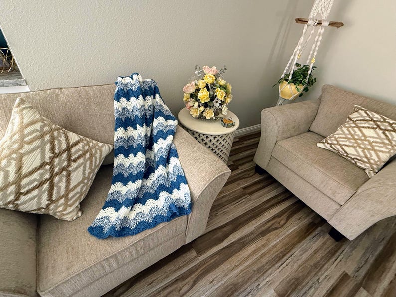 May include: Two beige armchairs with throw pillows, one with a blue and white crocheted throw blanket. The chairs are in a living room with a wooden floor and a small table with a bouquet of yellow and pink flowers.