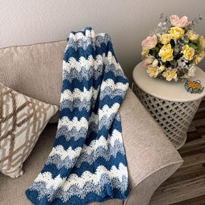 May include: A blue and white crocheted blanket with a wavy pattern, draped over a beige couch with a pillow. The blanket is made with a soft yarn and has a textured look.