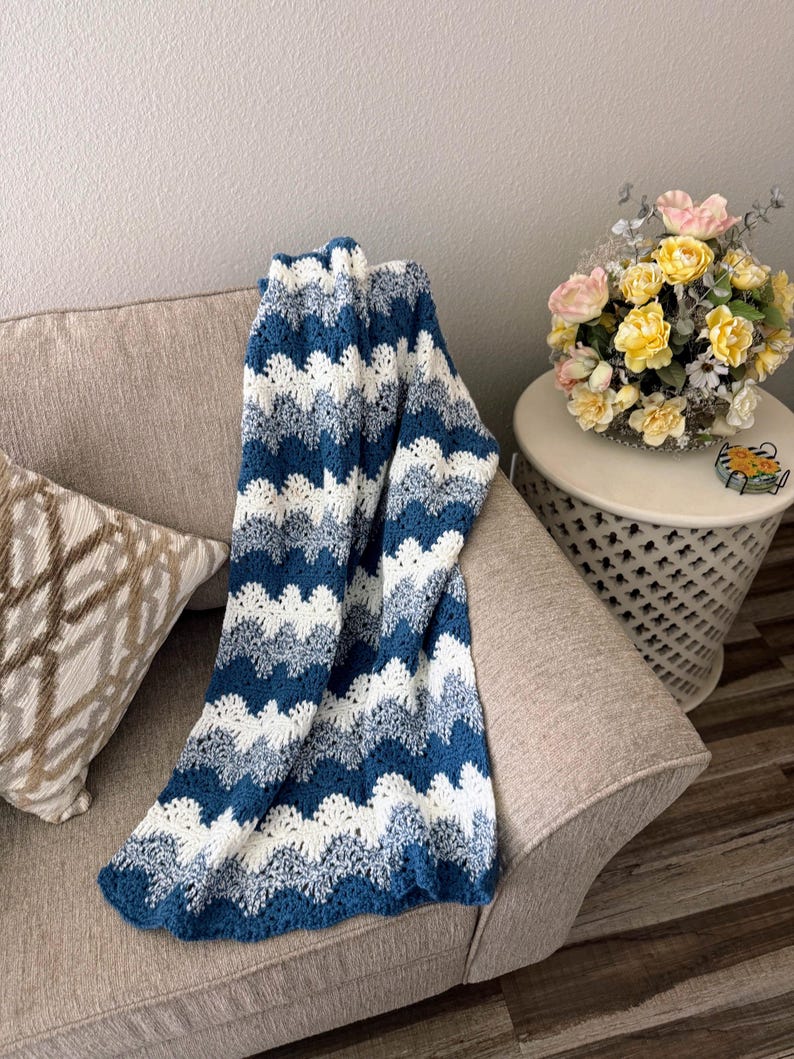 May include: A blue and white crocheted blanket with a wavy pattern, draped over a beige couch with a pillow. The blanket is made with a soft yarn and has a textured look.