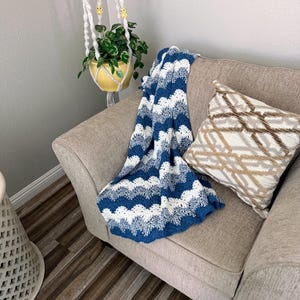 May include: A blue and white crocheted throw blanket with a wave pattern is draped over a light brown armchair. A brown and white patterned pillow is on the chair.