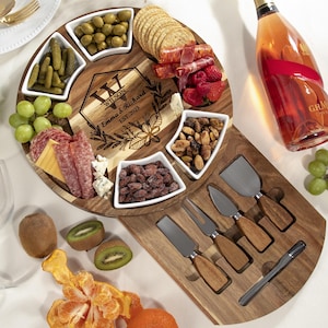 May include: A round wooden serving board with a built-in cheese knife holder. The board has six small white bowls for serving snacks like olives, crackers, cheese, and nuts. The board is engraved with the text "Wilson Emma & Richard Est. 2023".