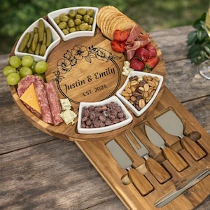 May include: A wooden charcuterie board with a floral design and the names "Justin & Emily EST. 2024" engraved. The board holds various snacks in white bowls, including olives, pickles, grapes, nuts, and dried fruit, along with crackers, cheese, and meats. Includes cheese knives.