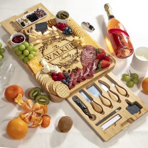 May include: A bamboo charcuterie board with the engraved text "The Walker Family" surrounded by a floral design. The board is filled with cheese, crackers, fruit, and meats. Includes a bottle of champagne and cheese knives.