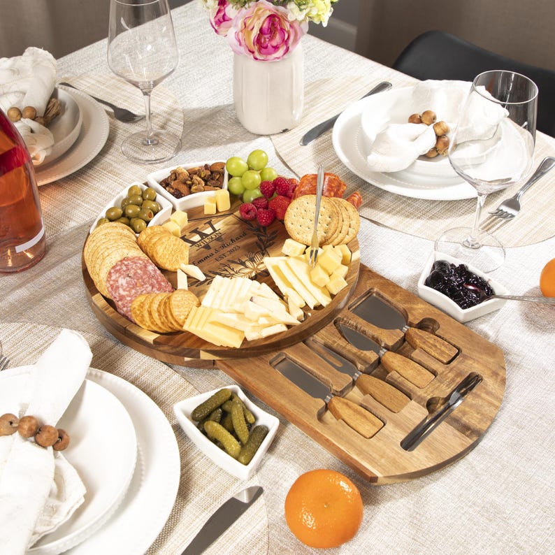 May include: A wooden cheese board with a variety of cheeses, crackers, salami, grapes, and berries. The board includes small white bowls with olives, nuts, and pickles. A set of cheese knives is included. The board is set on a table with wine glasses and plates.