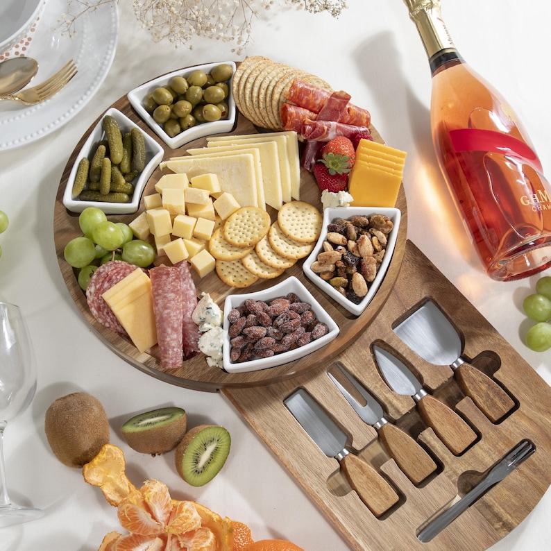 May include: A wooden charcuterie board with various cheeses, crackers, olives, pickles, salami, nuts, and grapes. A bottle of rose champagne and a set of cheese knives are also present. The board is arranged on a white surface.