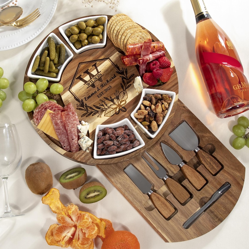 May include: A wooden charcuterie board with a personalized engraving, surrounded by small white bowls filled with olives, pickles, nuts, and other snacks. Includes cheese knives and a bottle of rose champagne.