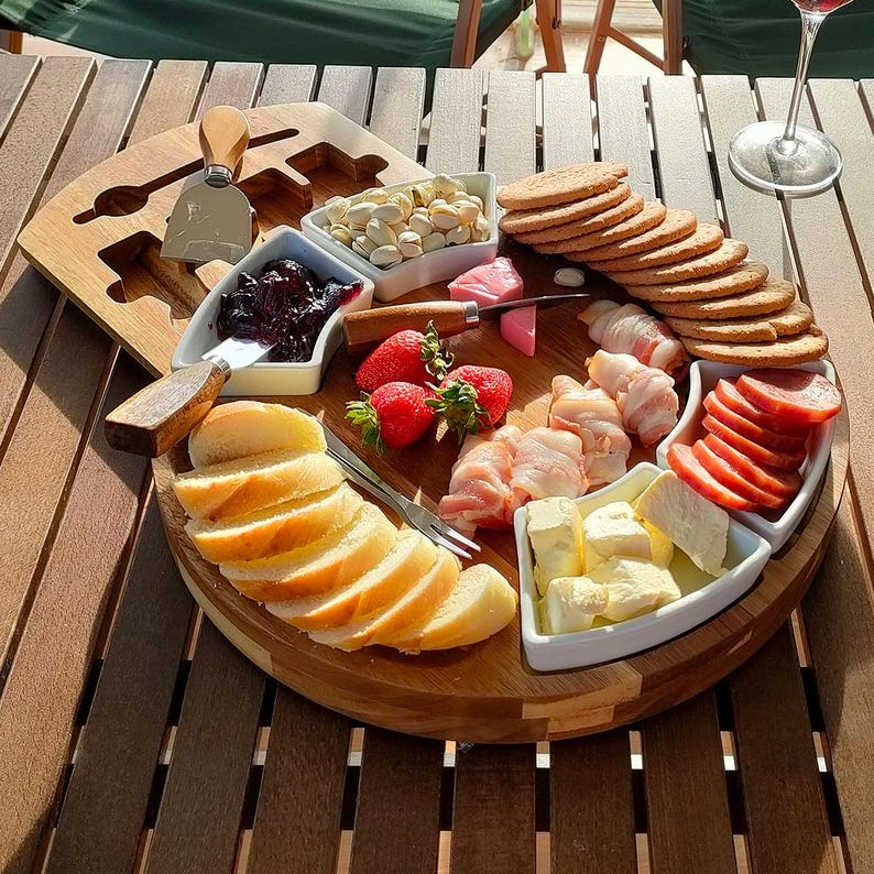 May include: A wooden charcuterie board with various food items arranged on it. Includes sliced bread, crackers, strawberries, cheese, meat, and nuts. A cheese knife and serving utensils are also present. The board is on a wooden table.